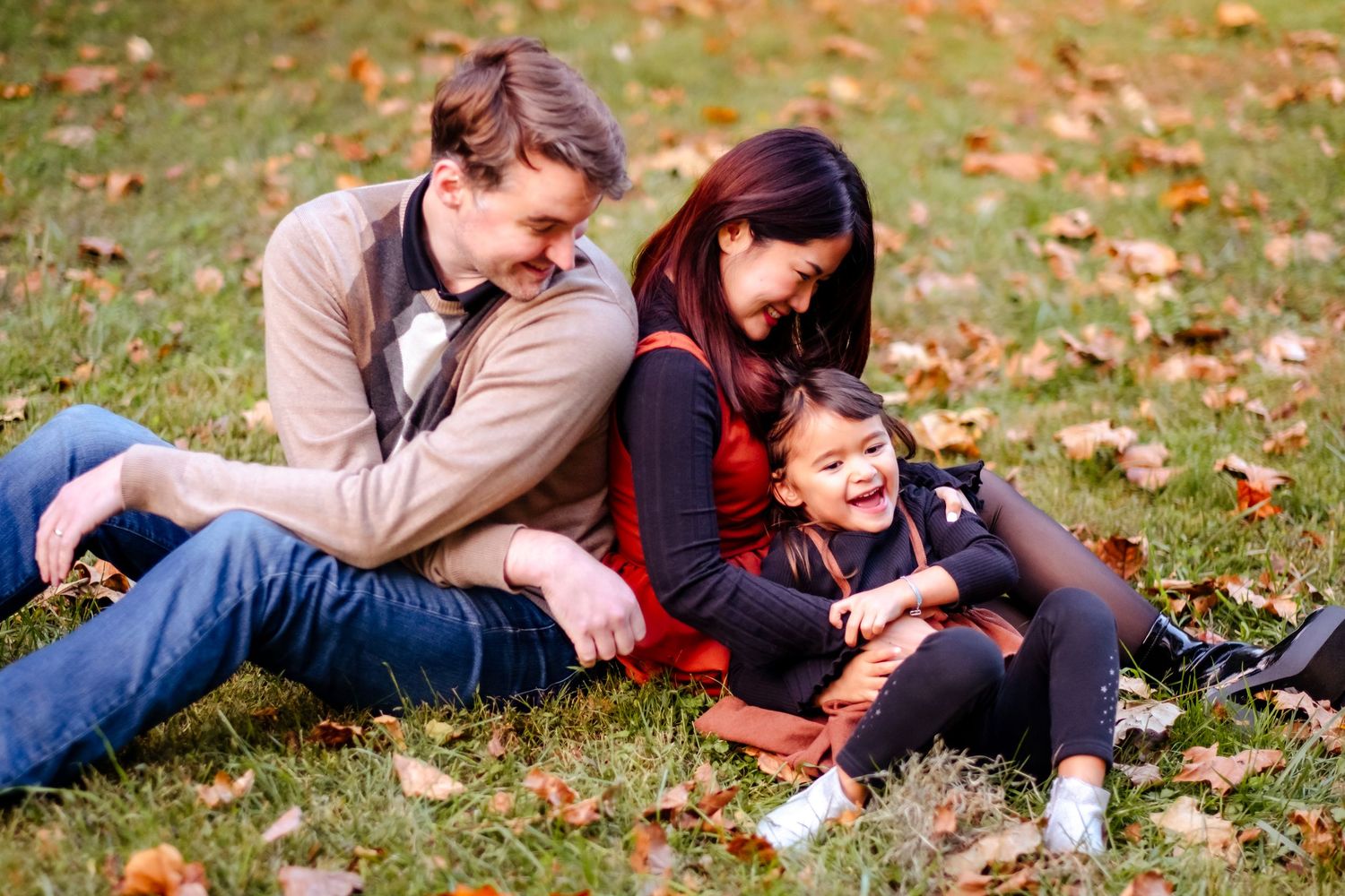 Family of three sitting on autumn grass, laughing together outdoors among fallen leaves.