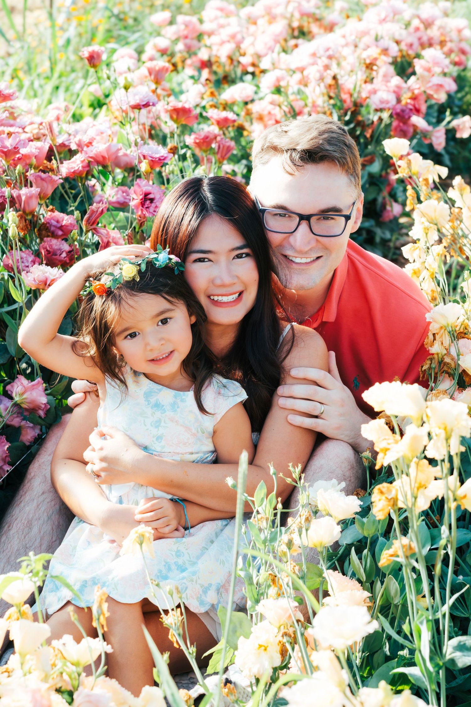 Happy interracial family with young daughter sitting in colorful flower garden, smiling together outdoors.