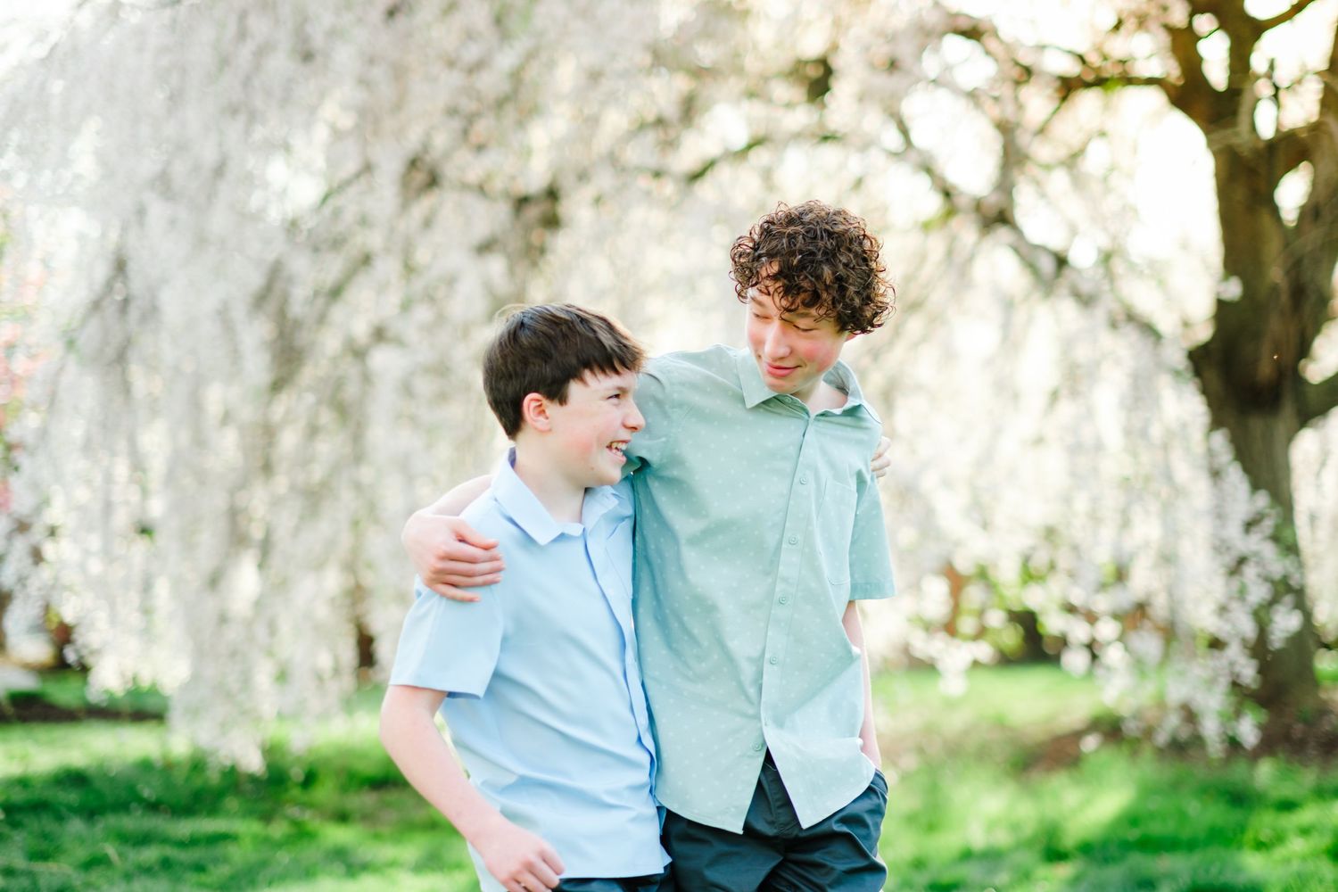 Two young men smiling and embracing outdoors under blooming white cherry blossom trees in spring.