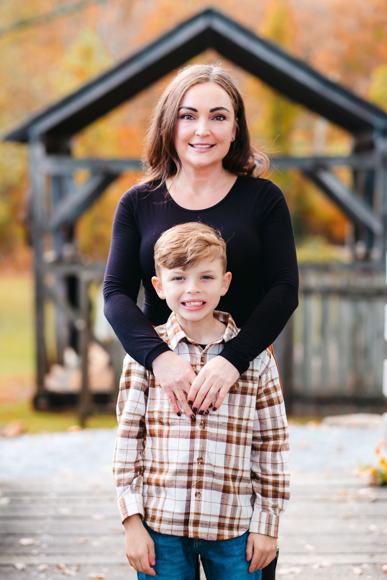 A woman and young boy pose together outdoors in fall, she wears black top, he wears plaid shirt, colorful autumn trees behind.