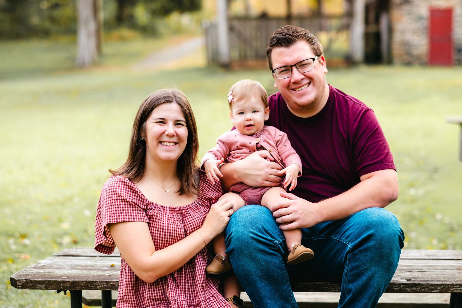 Happy family of three sitting on a bench outdoors, parents smiling with baby in red outfits on a green lawn.