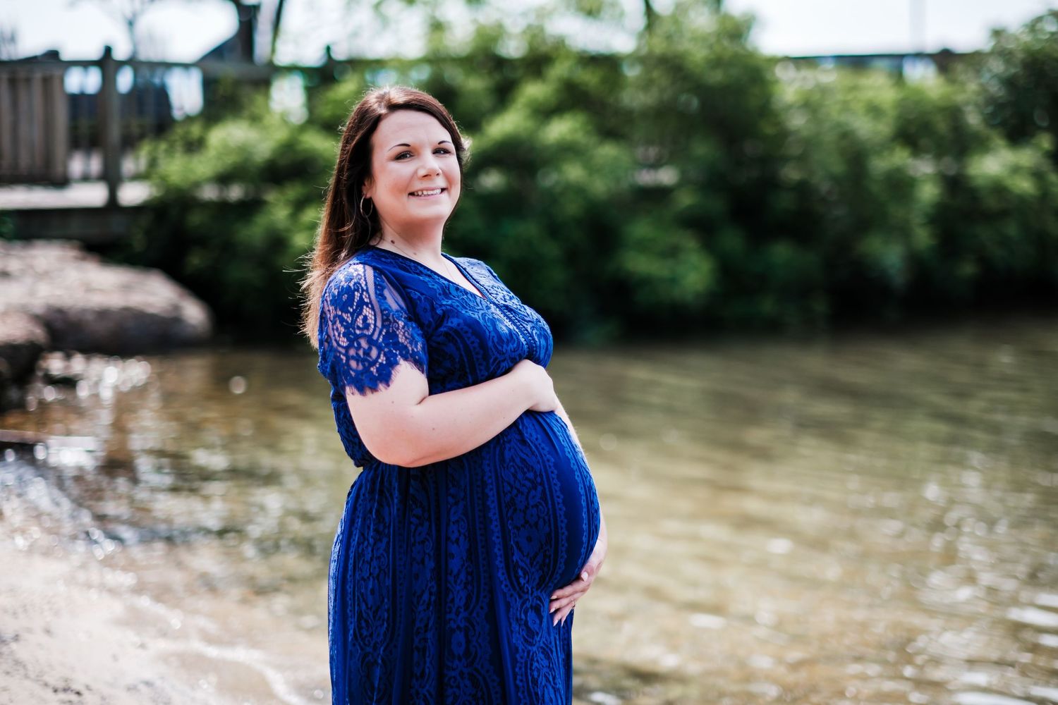 Pregnant woman in blue lace maternity dress posing by a river, cradling her baby bump outdoors.