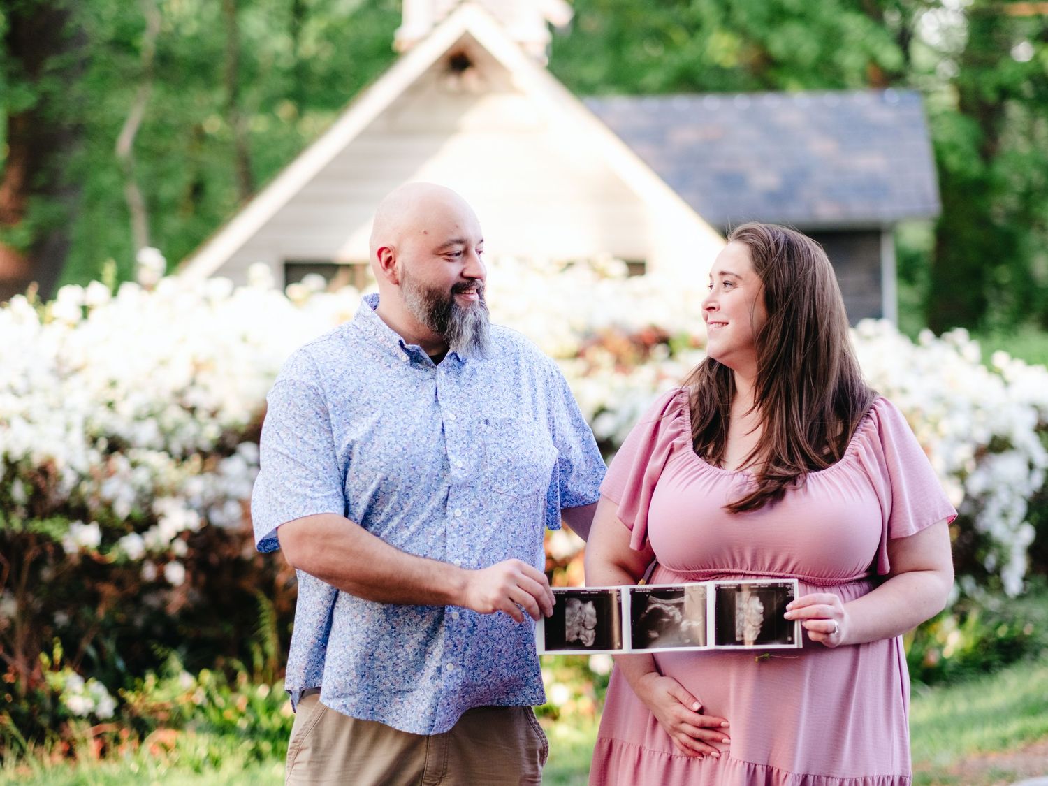 Happy couple smiling outdoors holding ultrasound photos, announcing pregnancy near a white cottage with blooming flowers.