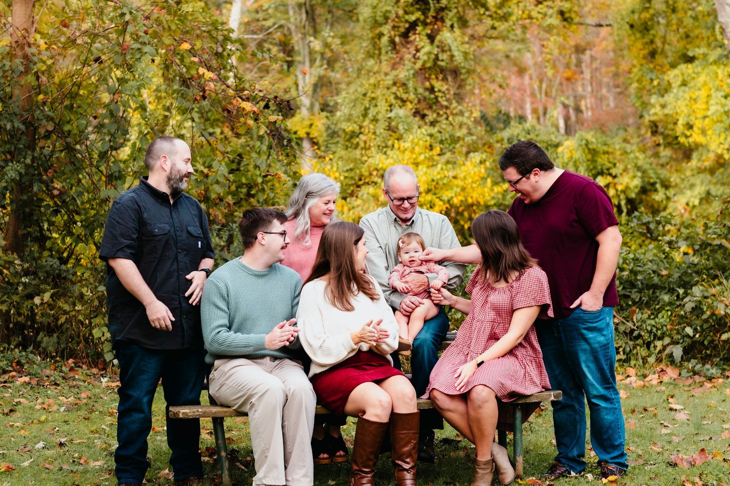 Multi-generational family gathering outdoors in autumn, smiling together on a bench surrounded by colorful fall foliage.