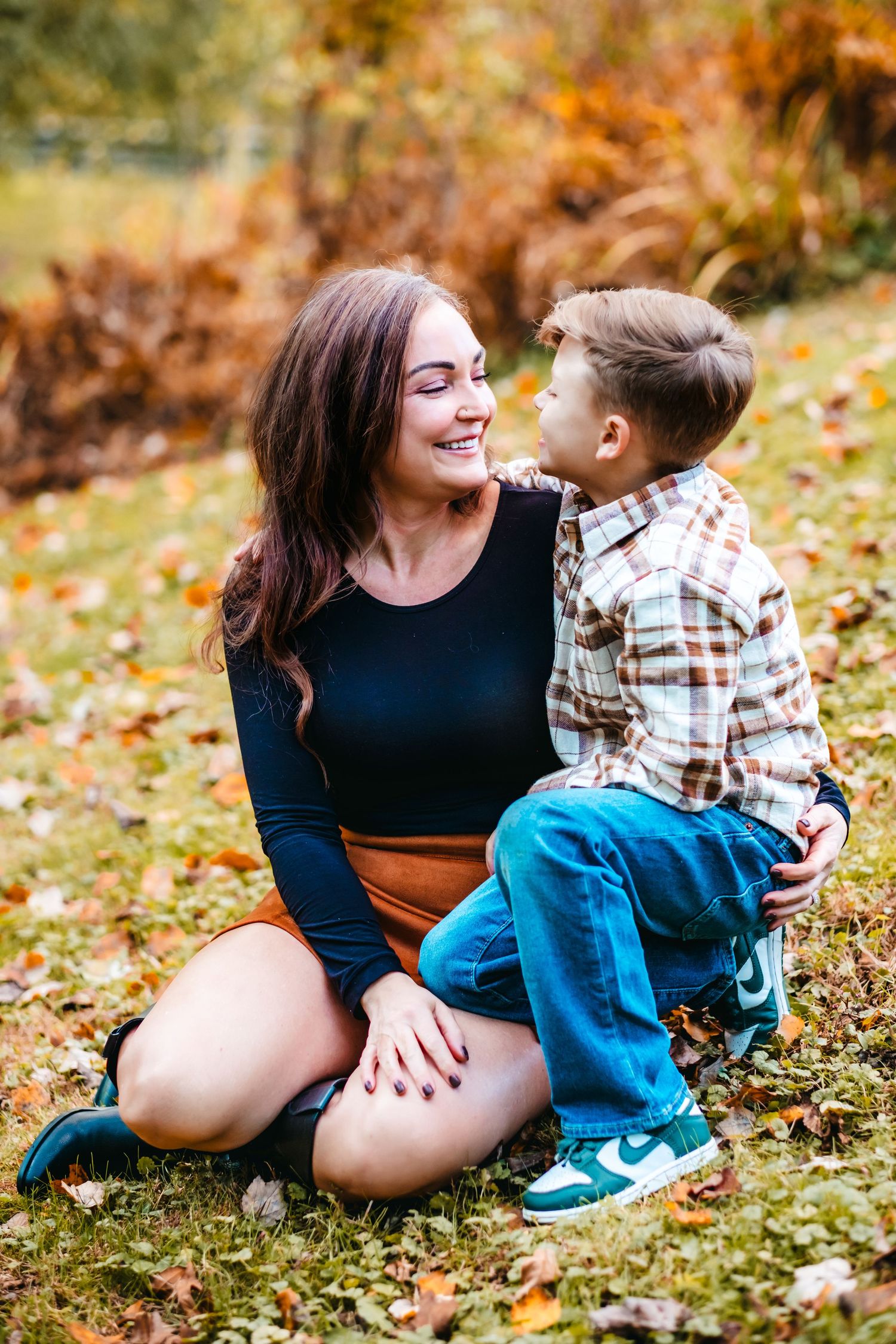 A smiling mother sits on autumn grass with her young son in a plaid shirt, sharing a tender moment outdoors.