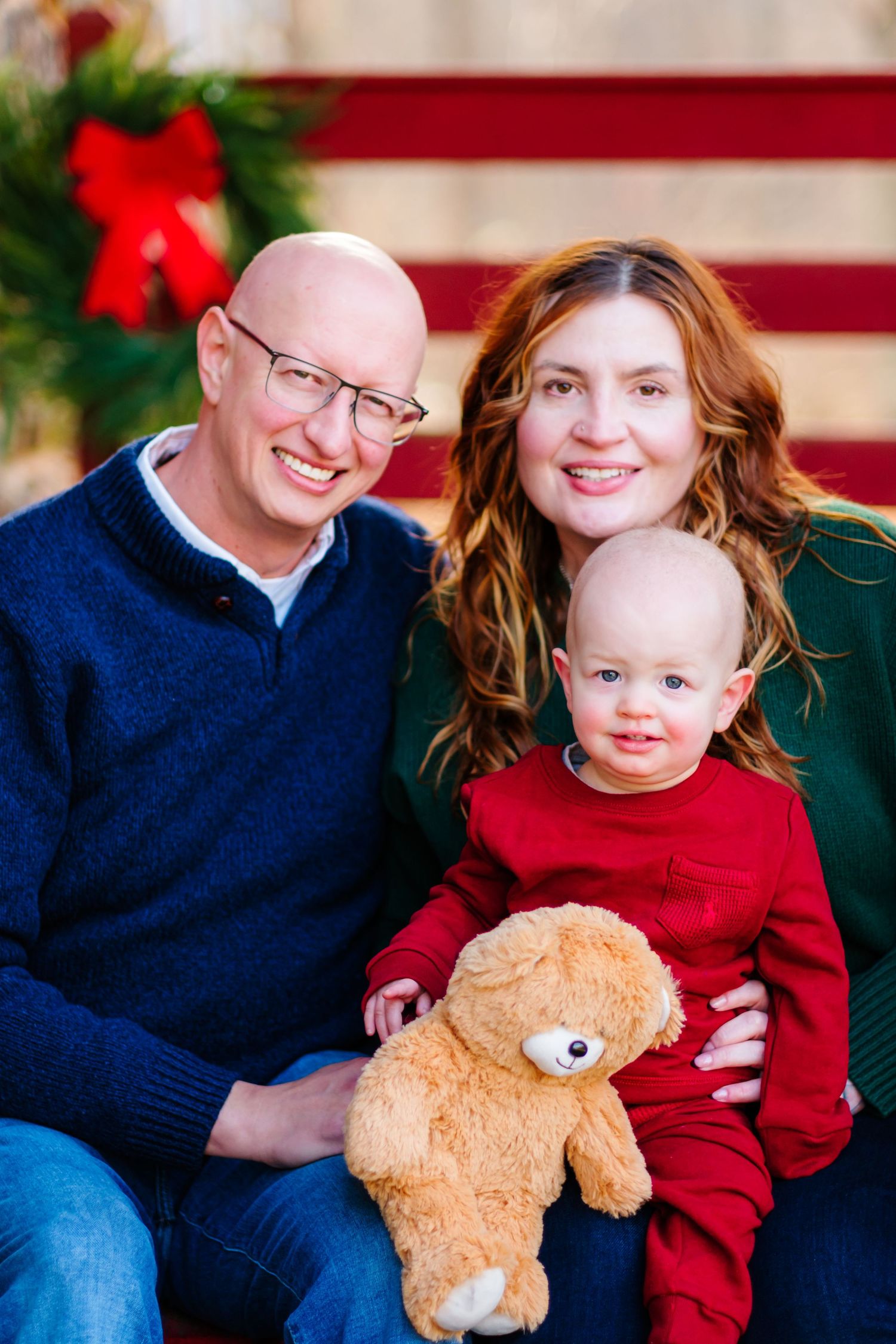A smiling family of three poses for a Christmas portrait, with a bald child holding a teddy bear between his parents.