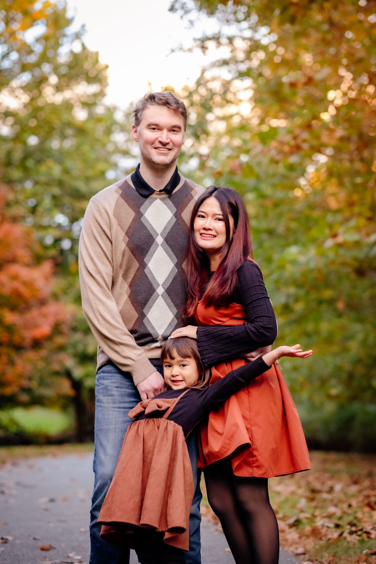 Family of three posing outdoors in autumn park, parents smiling with young daughter in matching rust-colored outfits.