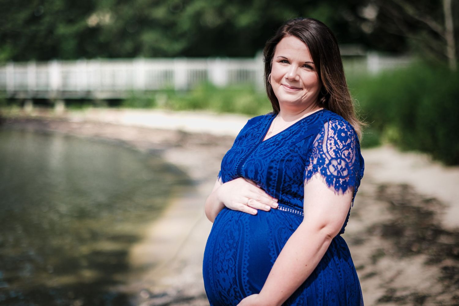 Pregnant woman in blue lace dress posing outdoors near a serene river, smiling warmly during a maternity photo session.