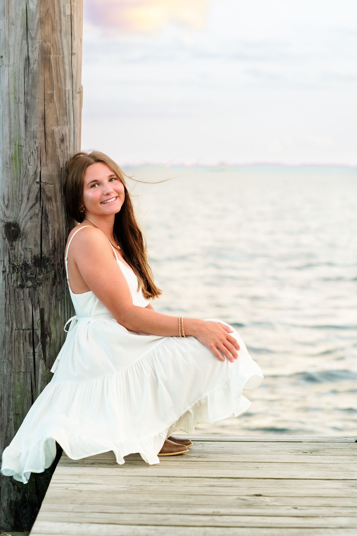 Young woman in white dress sitting on a wooden dock by the ocean at sunset.