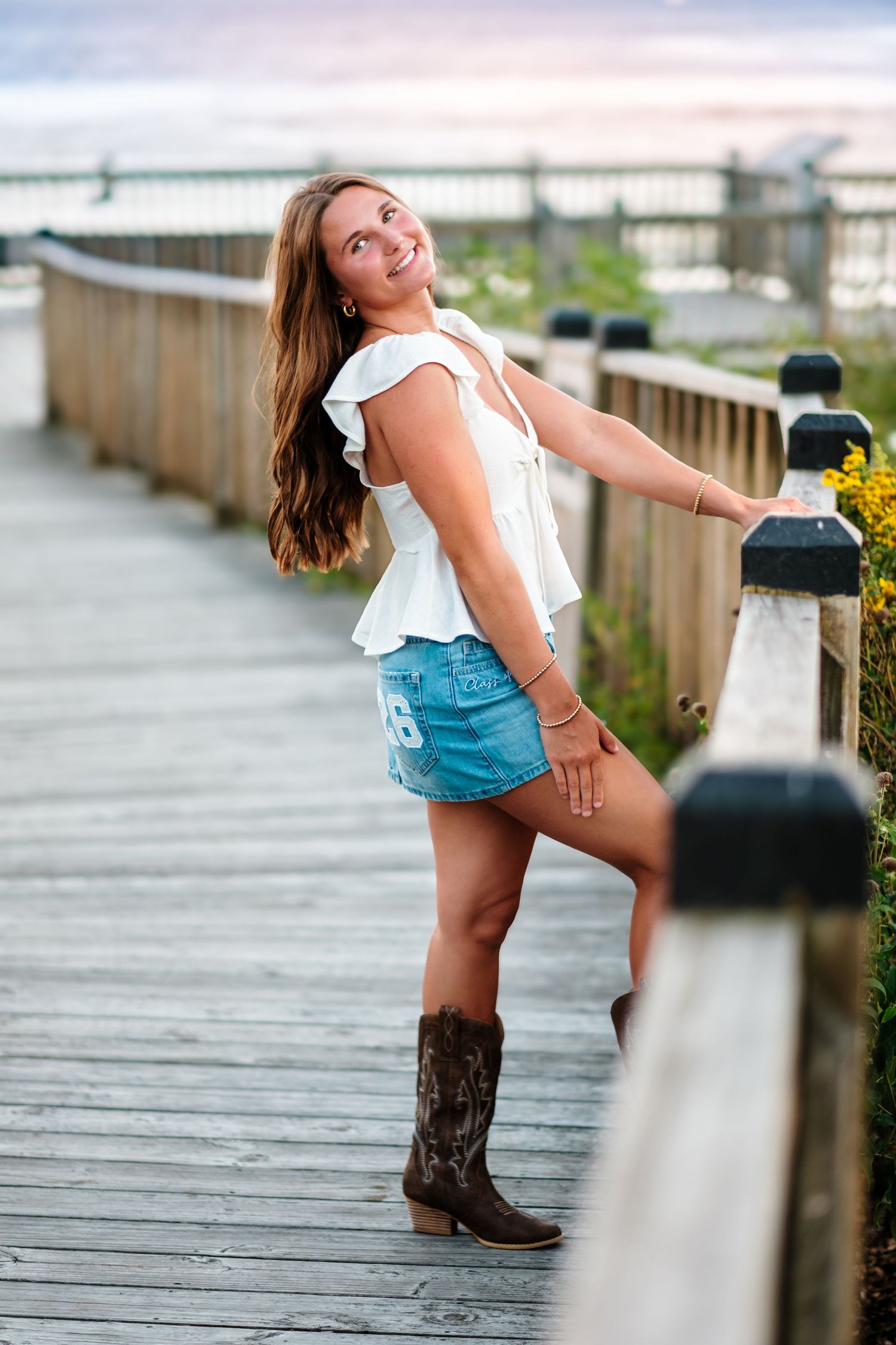Young woman in white top, denim skirt, and cowboy boots posing on a wooden boardwalk at sunset.
