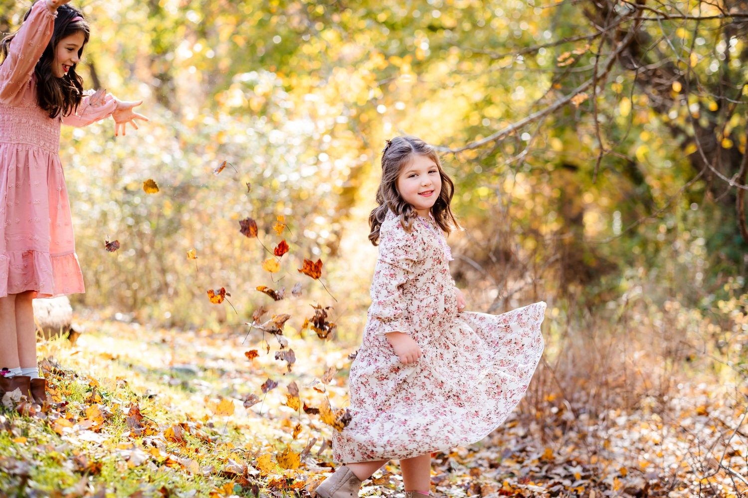 Two girls in floral dresses twirl and toss autumn leaves in a sunlit forest during fall.