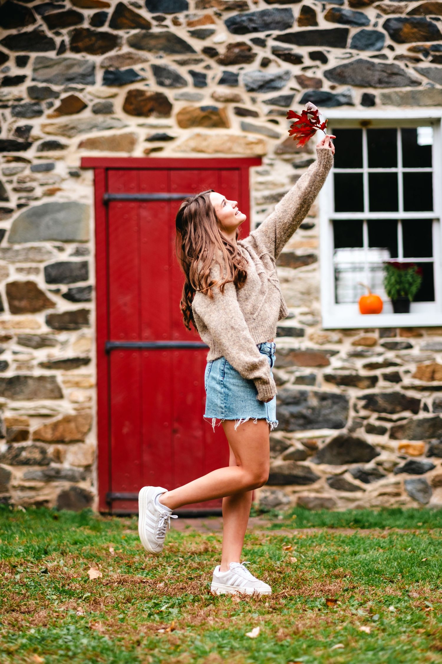 Young woman in sweater and denim skirt holding autumn leaf outside rustic stone building with red door in fall.