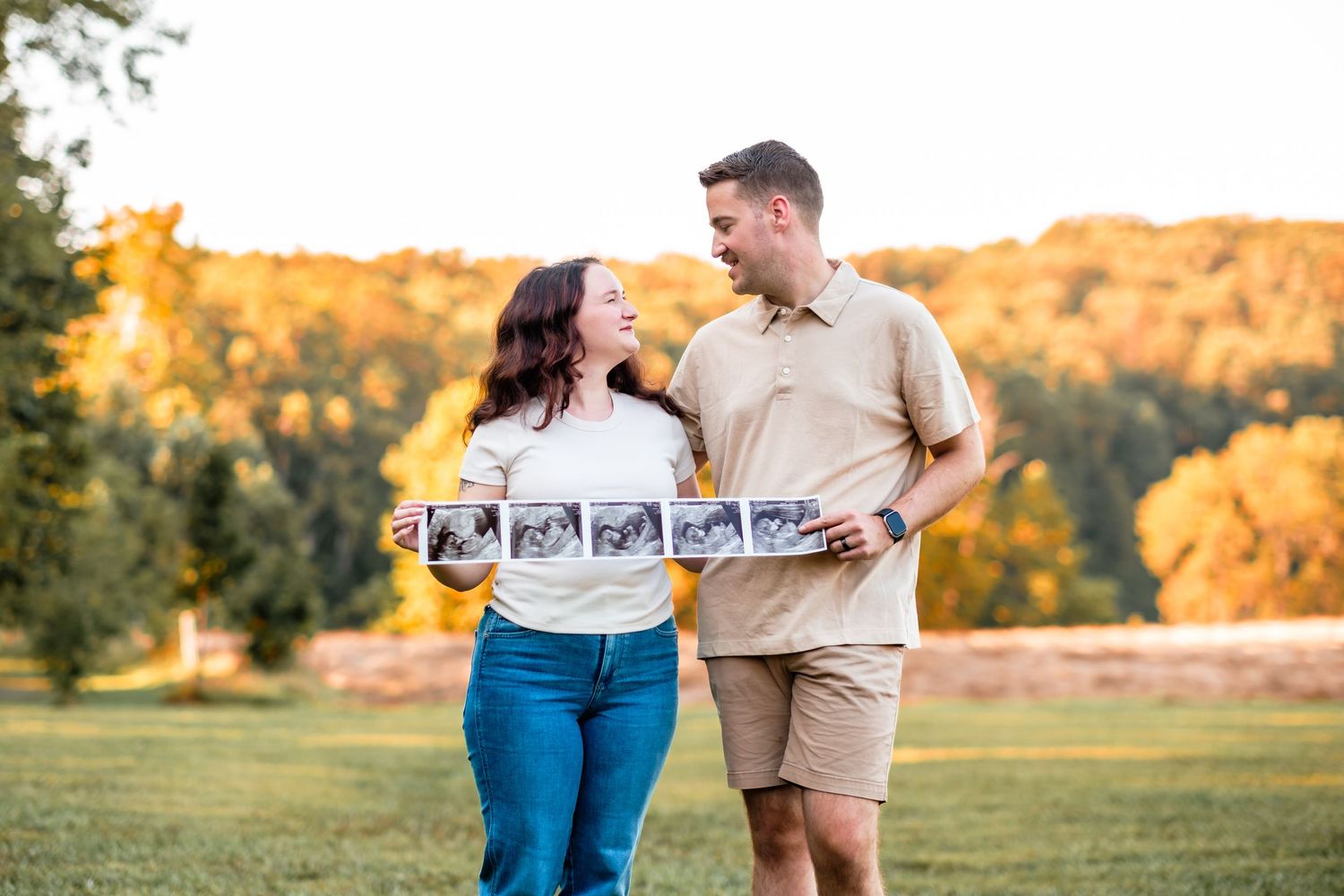 Couple smiling at each other outdoors holding ultrasound photos during a fall maternity announcement photoshoot.