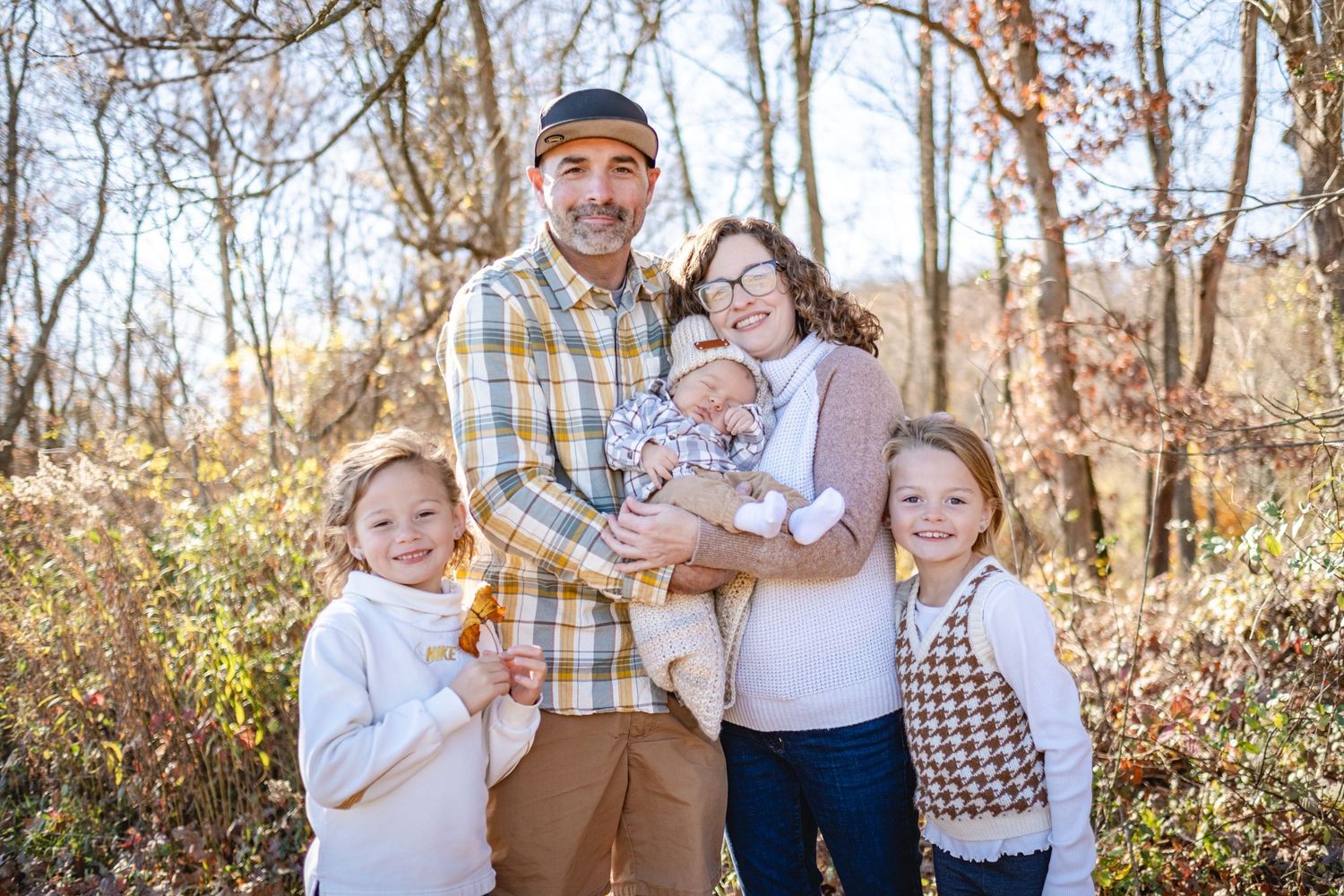 Family of five posing outdoors in autumn, parents holding newborn baby with two young girls smiling in fall foliage setting.