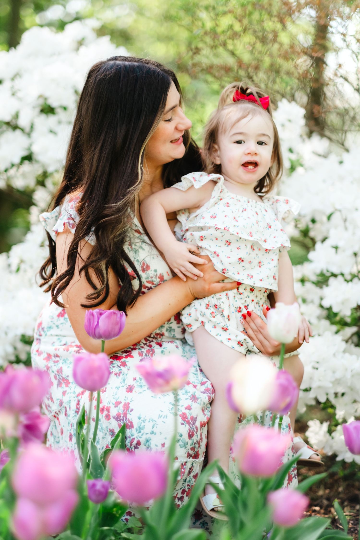A mother and toddler in matching floral dresses pose among pink tulips and white blooms in a spring garden.