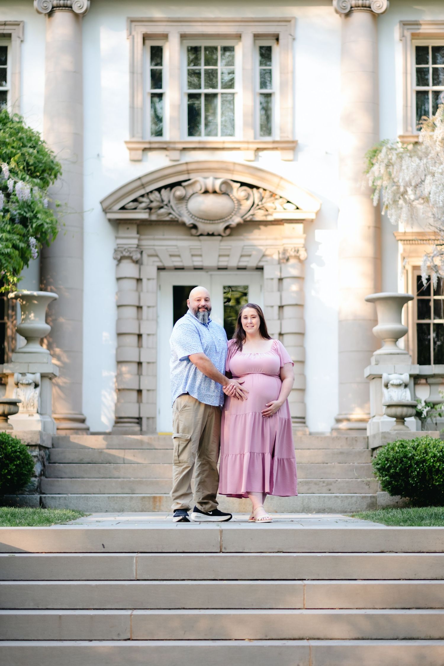 Couple poses on grand estate steps, pregnant woman in pink dress held by partner in blue shirt, elegant white mansion backdrop.