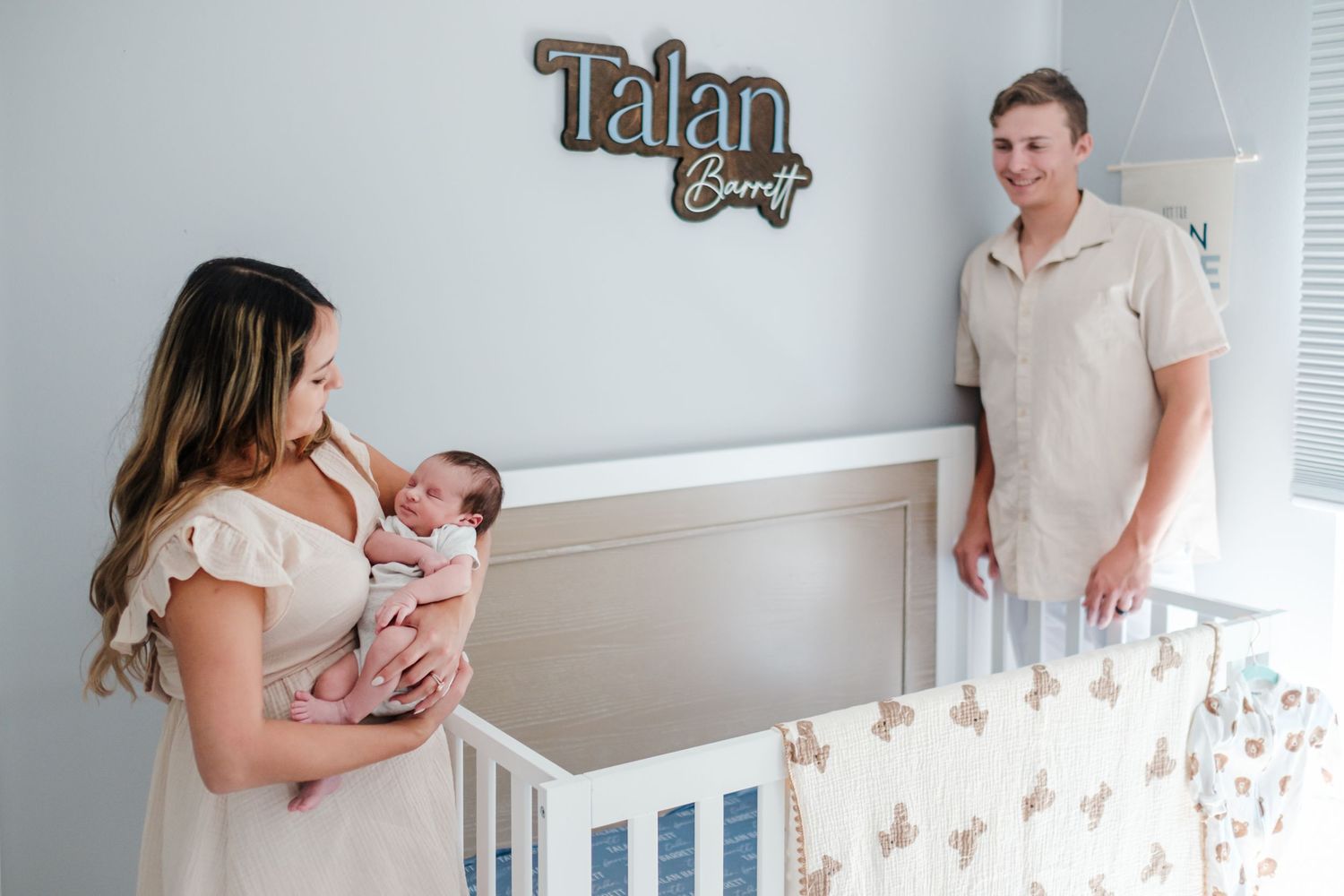 New parents stand in nursery with newborn baby near crib under Talan Barrett name sign on wall.