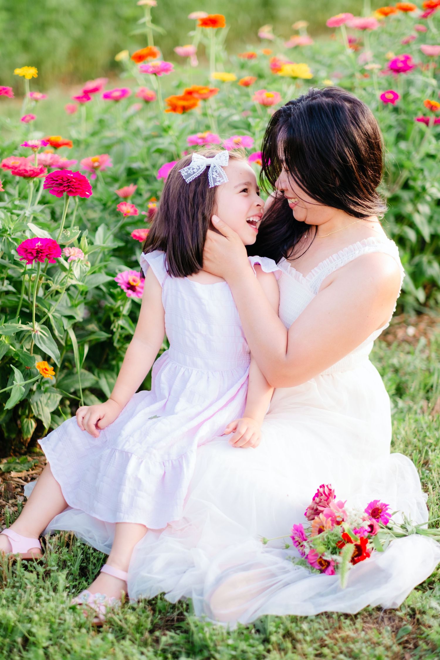 Mother and daughter in white dresses share a tender moment among colorful zinnia flowers in a garden.