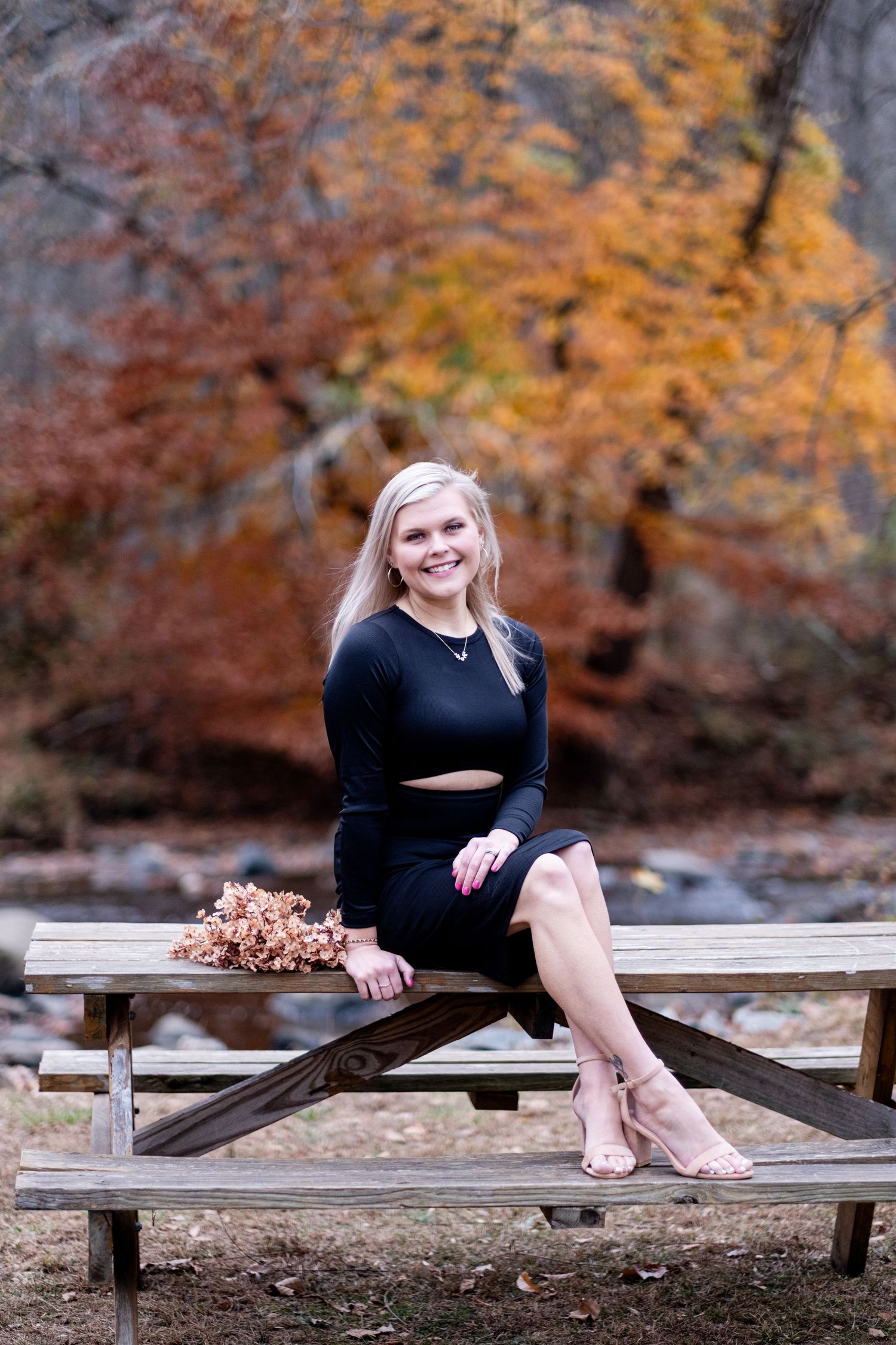 Smiling blonde woman in black dress sitting on picnic table with fall foliage backdrop.