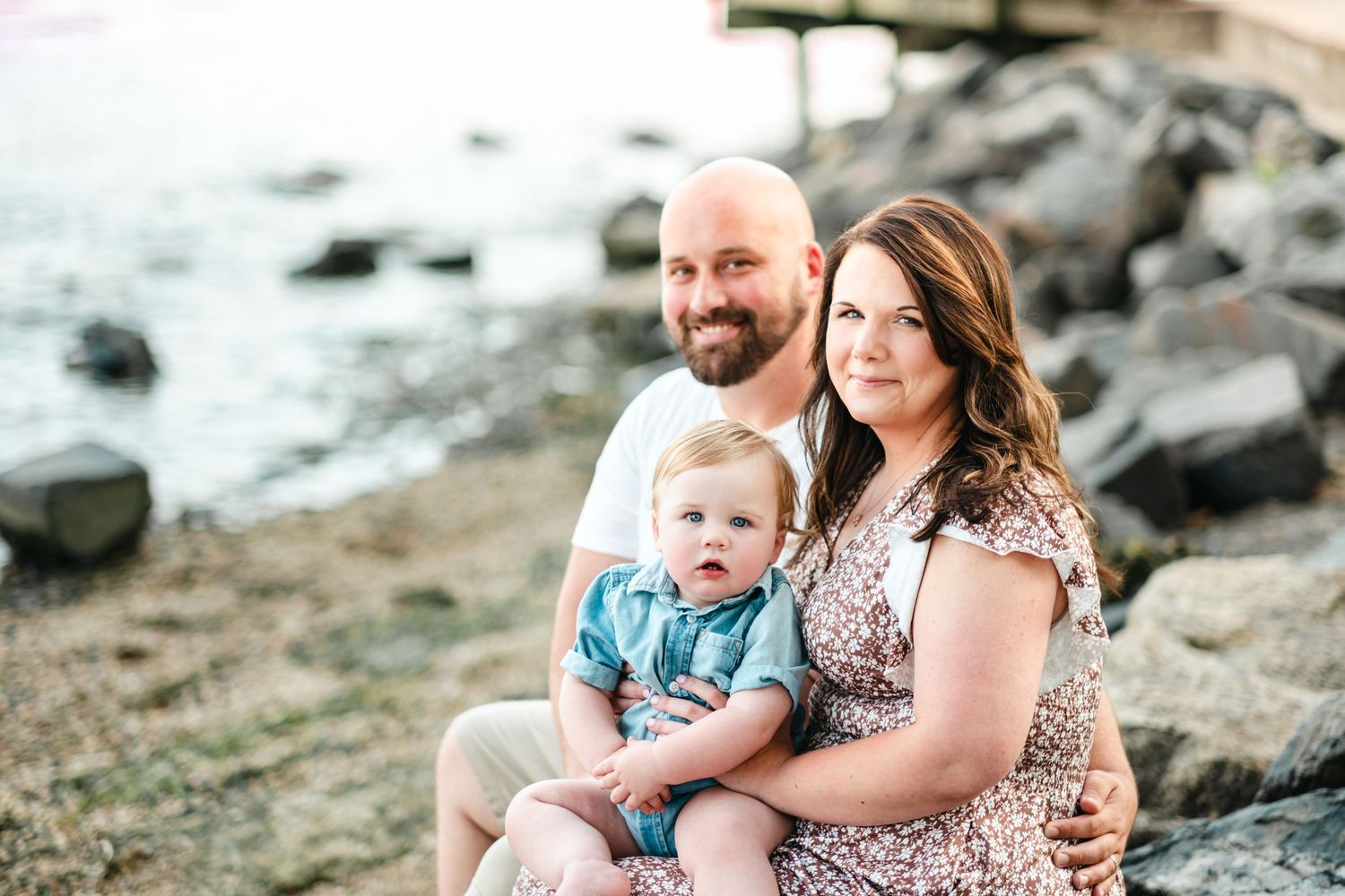 A happy family of three poses on rocky shoreline, with parents holding their baby boy in a floral dress and white shirt.