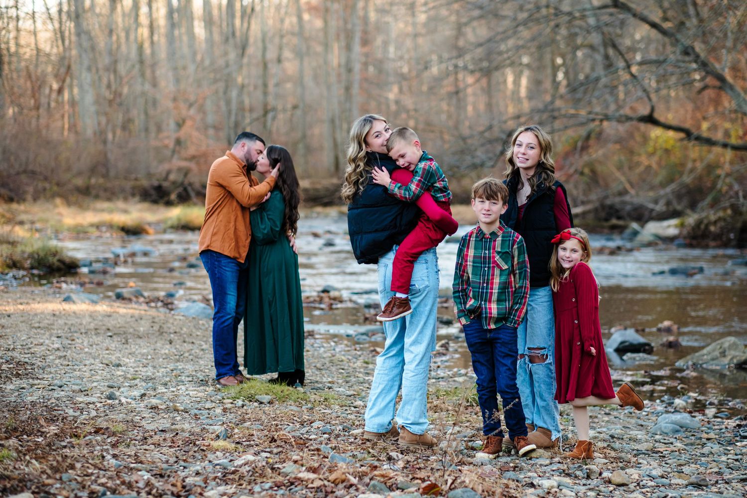 Family portrait outdoors by a creek in autumn, parents with children dressed in festive red, green, and blue outfits.