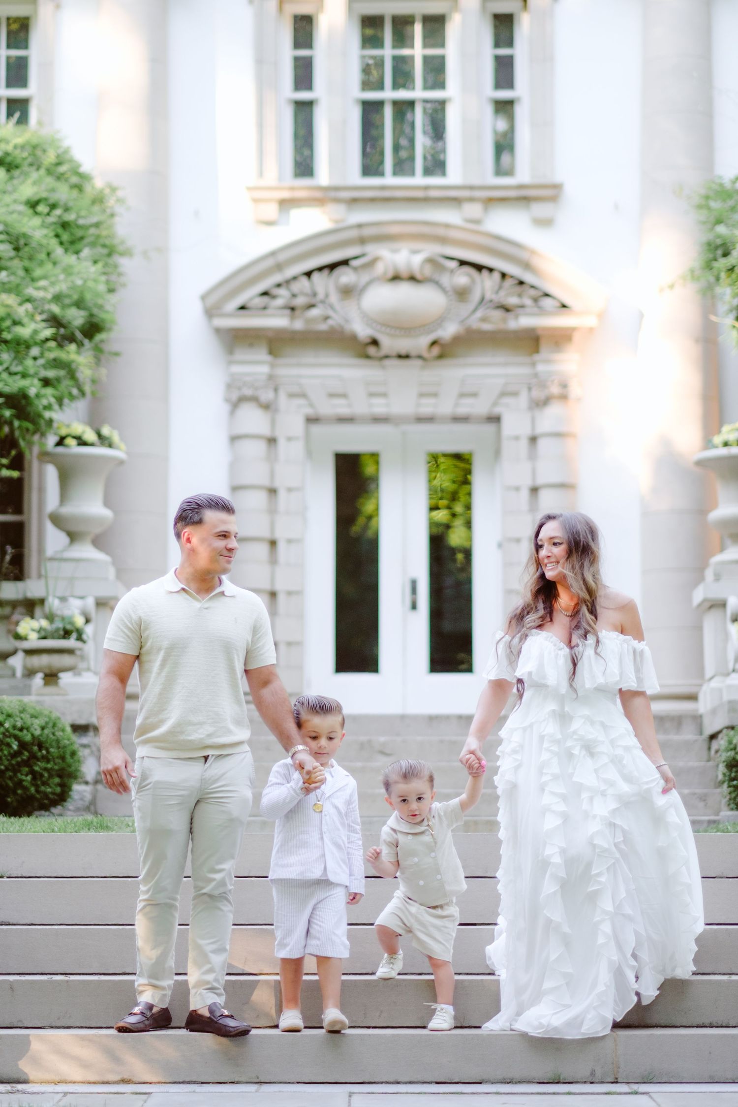 Family of four in white and beige outfits posing on steps of elegant mansion with ornate architecture.