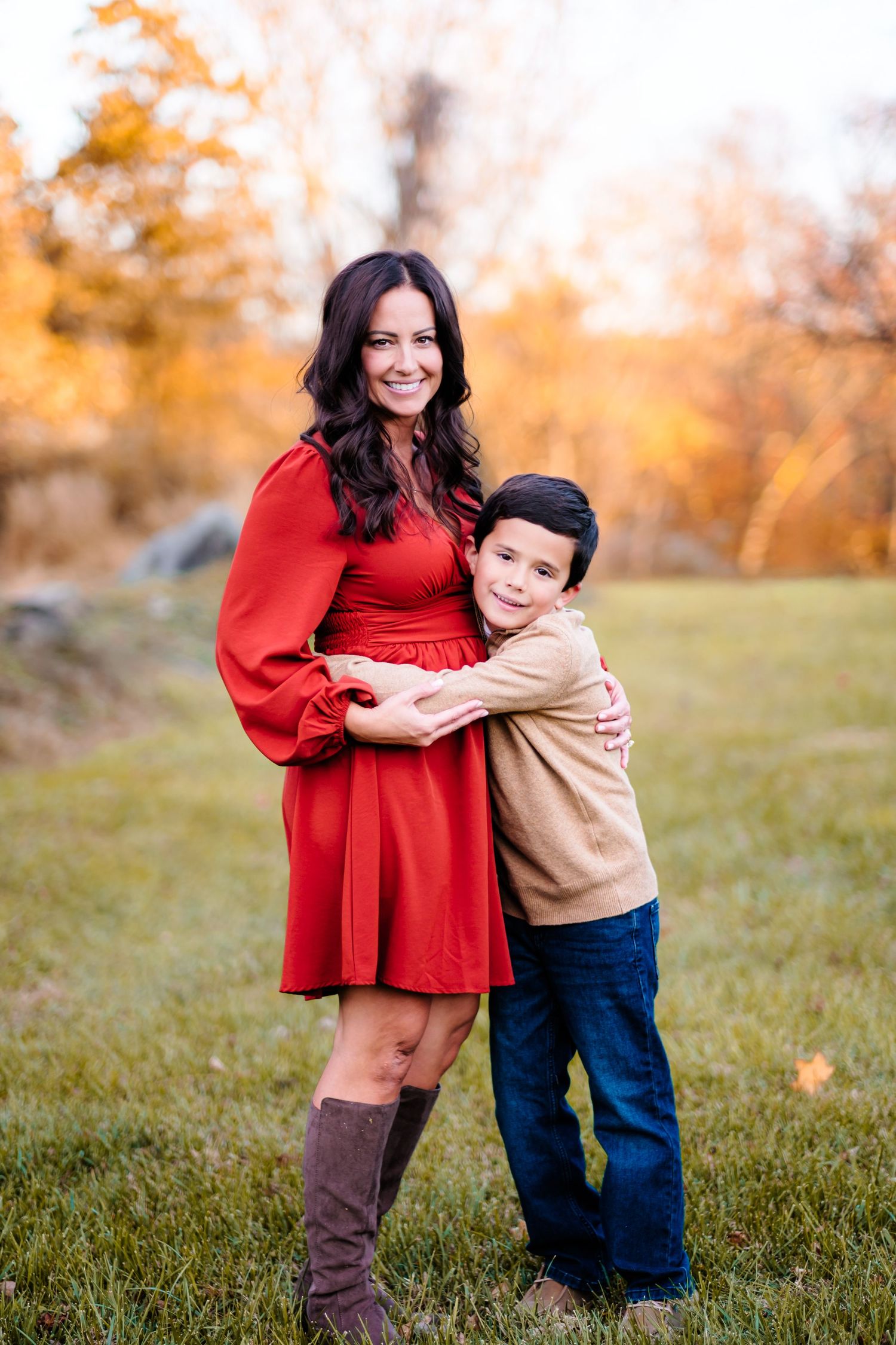 Mother in red dress and brown boots hugging her son in a colorful autumn park.