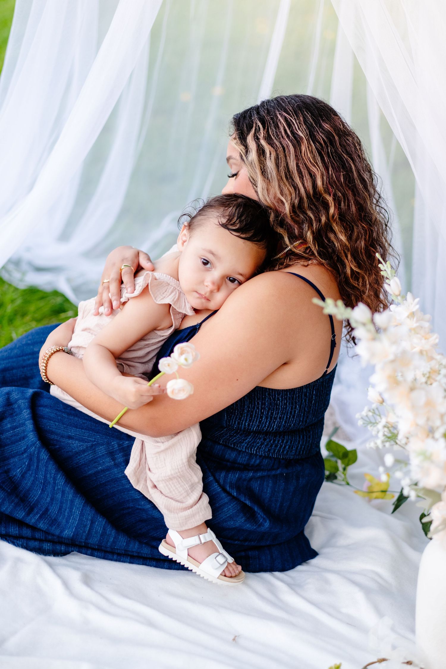 A mother in a blue dress tenderly embraces her baby girl outdoors under a white canopy with flowers.