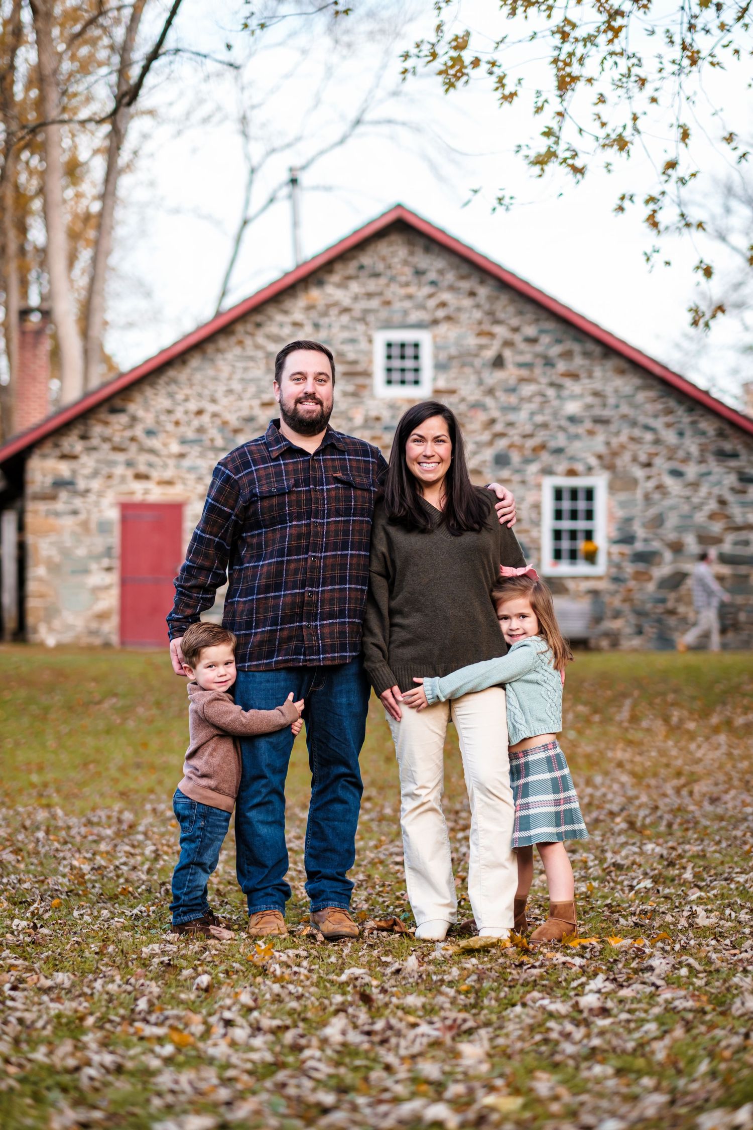 Family of four smiling outdoors in fall, standing before a rustic stone building with red trim.