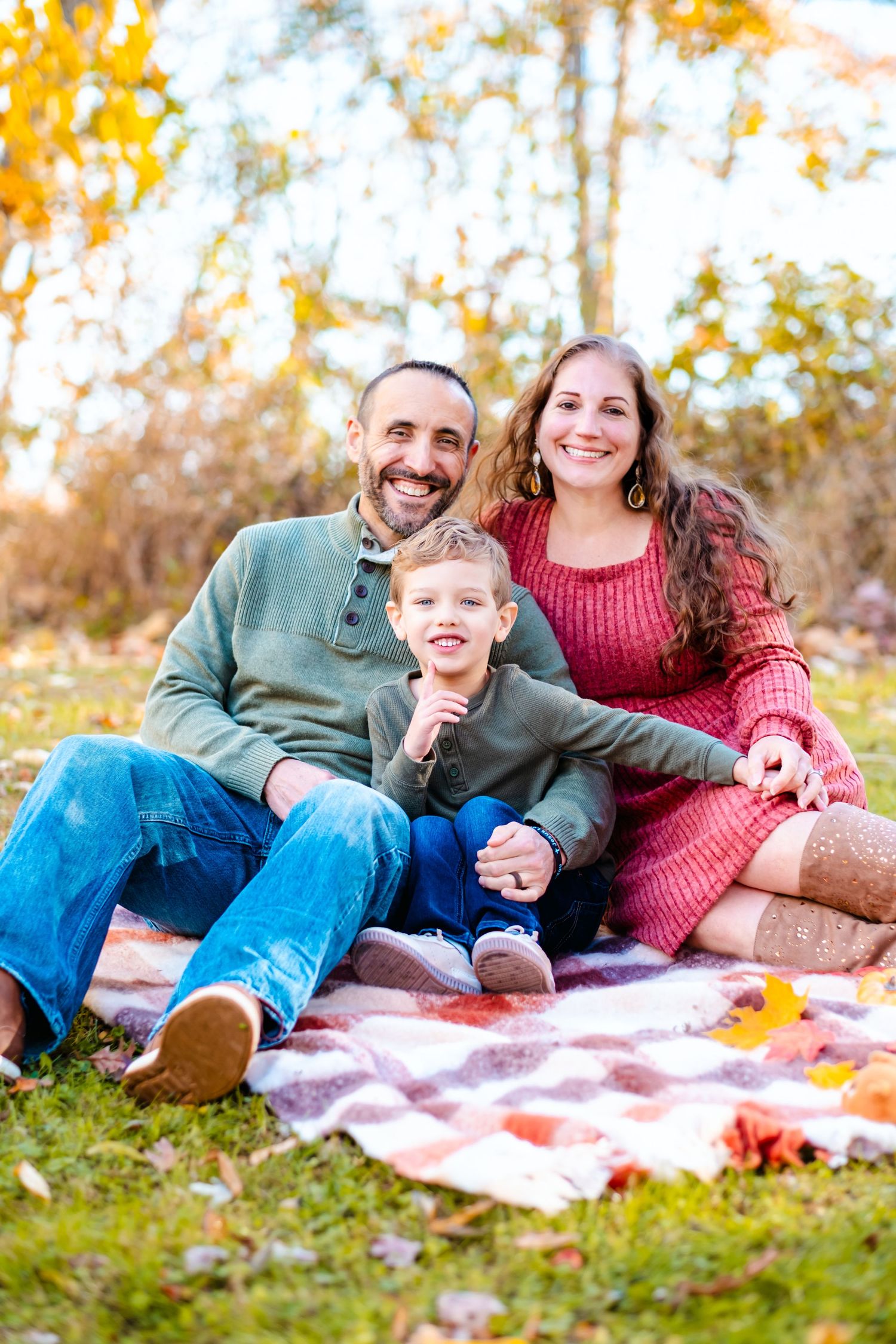 Family of three sitting on plaid blanket in autumn park, smiling parents with young boy among fall foliage.