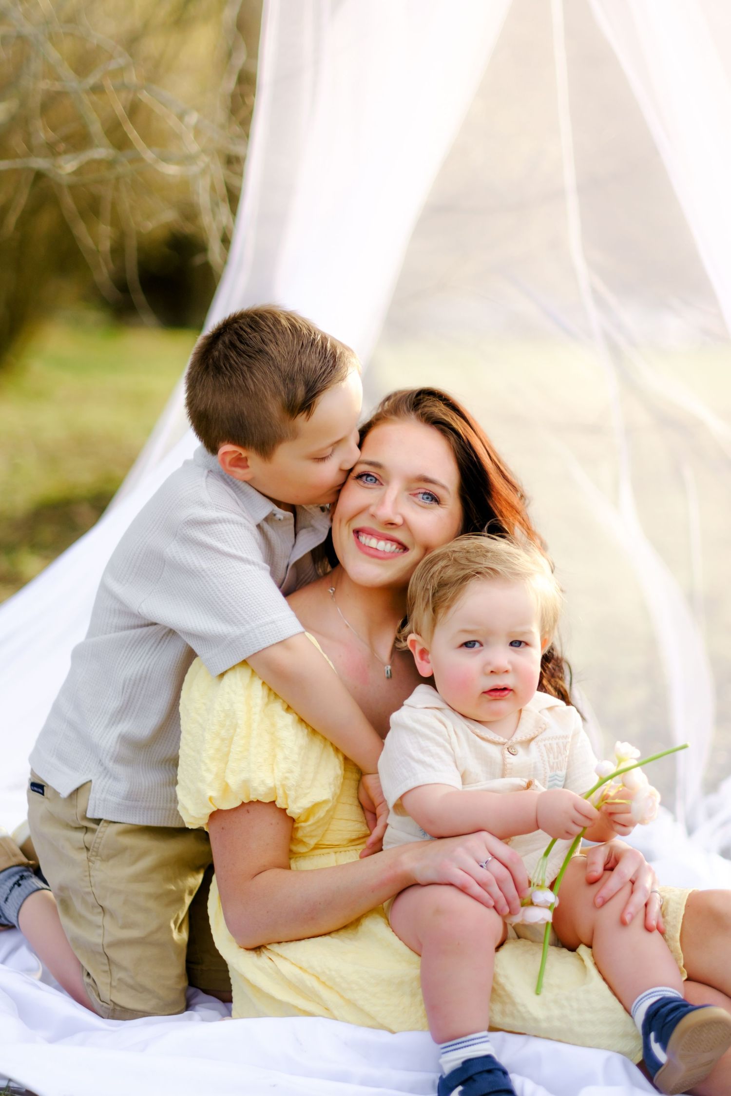 Smiling mother in yellow dress receives kiss from older son while holding toddler during outdoor family photo.