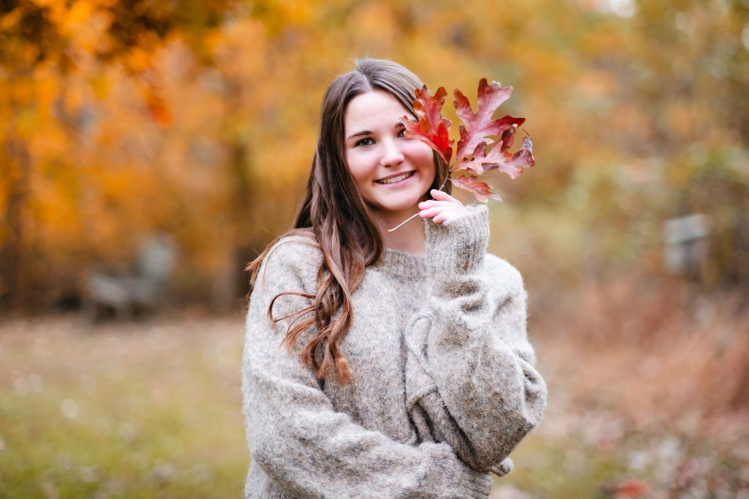 Young woman in cozy sweater holding vibrant red autumn leaves, smiling outdoors with colorful fall foliage background.