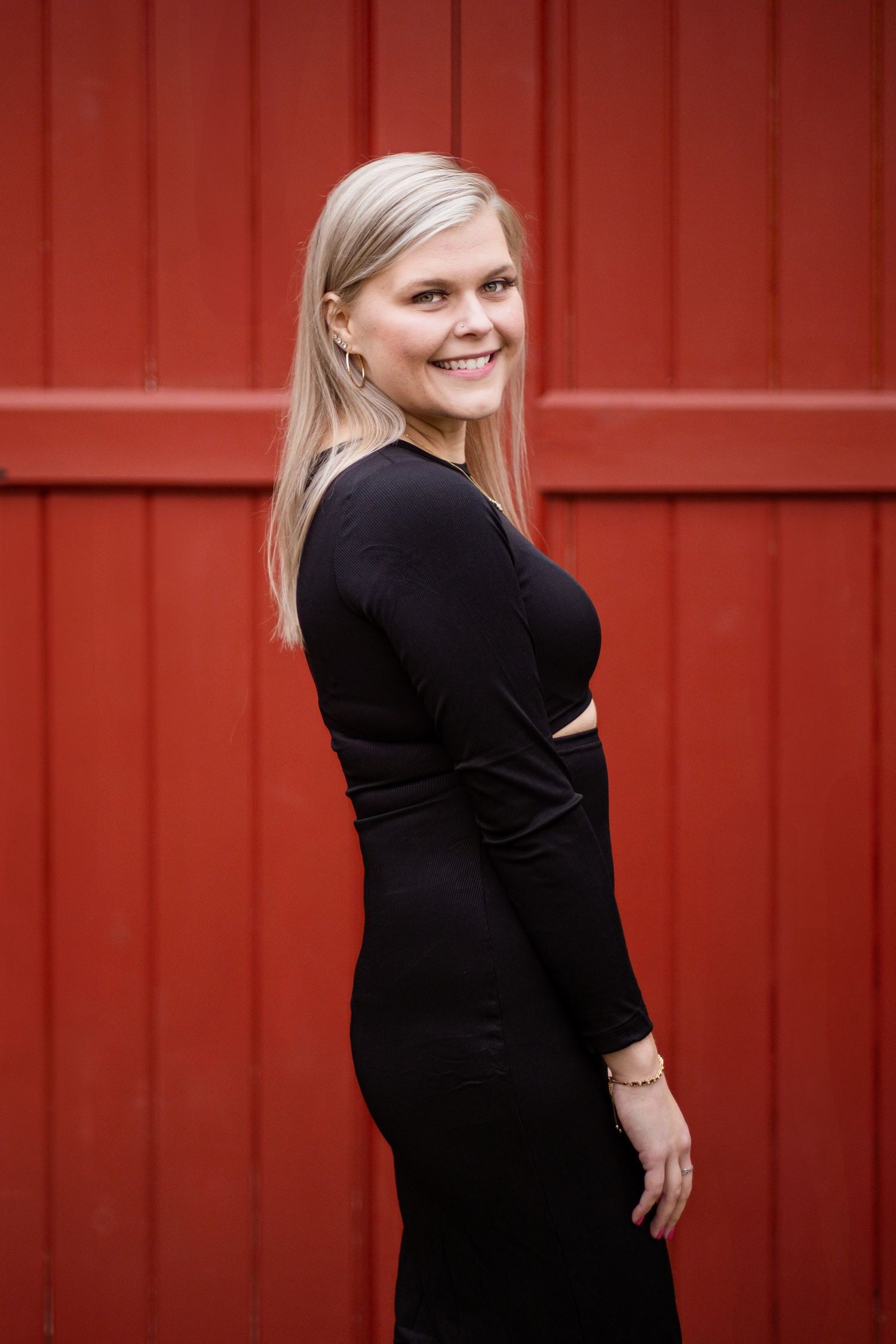 Smiling blonde woman in black long-sleeve dress posing against a red wooden wall background.