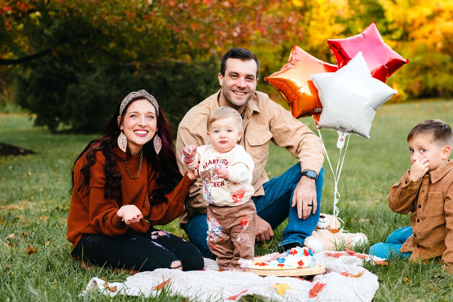 Family of four celebrating a first birthday outdoors on a blanket with star balloons and cake in an autumn park.
