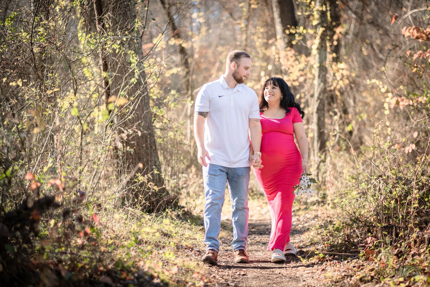 Pregnant couple holding hands walking on a sunlit forest path, woman in pink dress, man in white shirt and jeans.