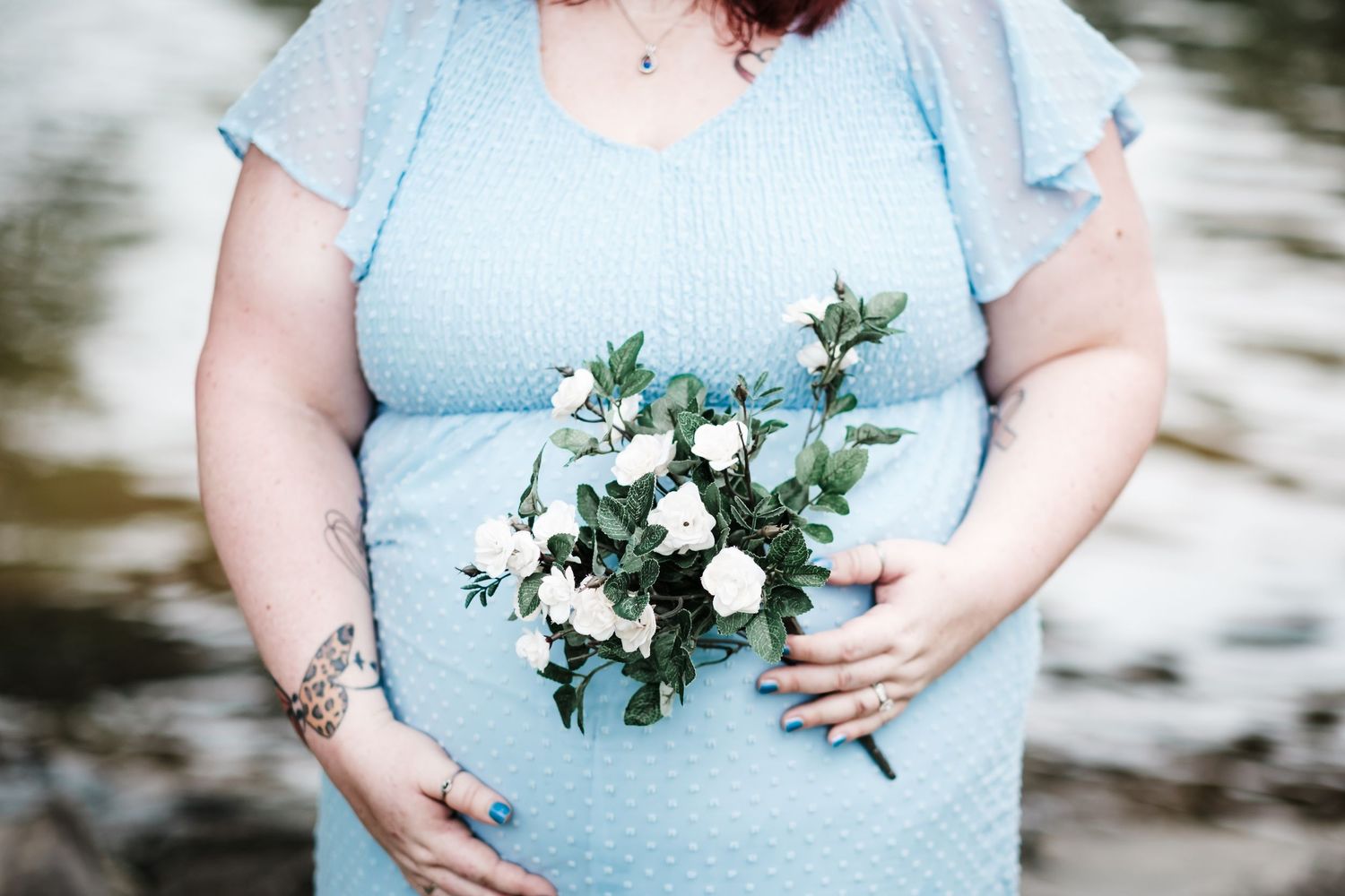 Pregnant woman in blue polka dot dress holding white flowers bouquet, showing tattoos on arms near a river.