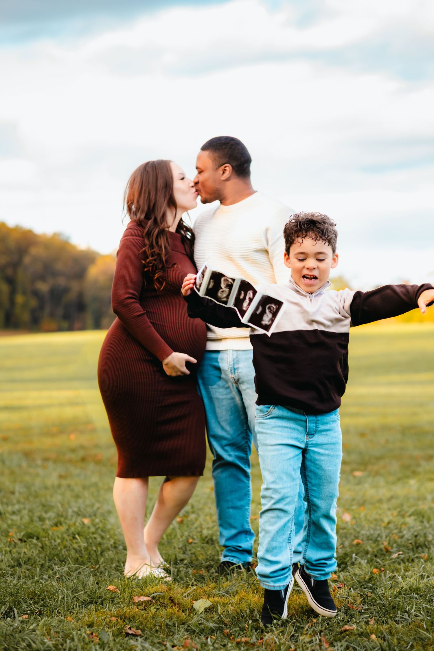 Pregnant woman and man kissing outdoors while excited boy holds ultrasound photos during fall maternity photo session.