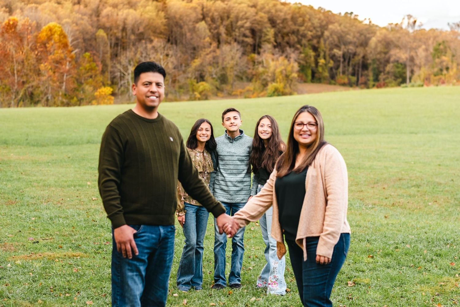 Family of five posing together in a green field with colorful autumn trees in the background.