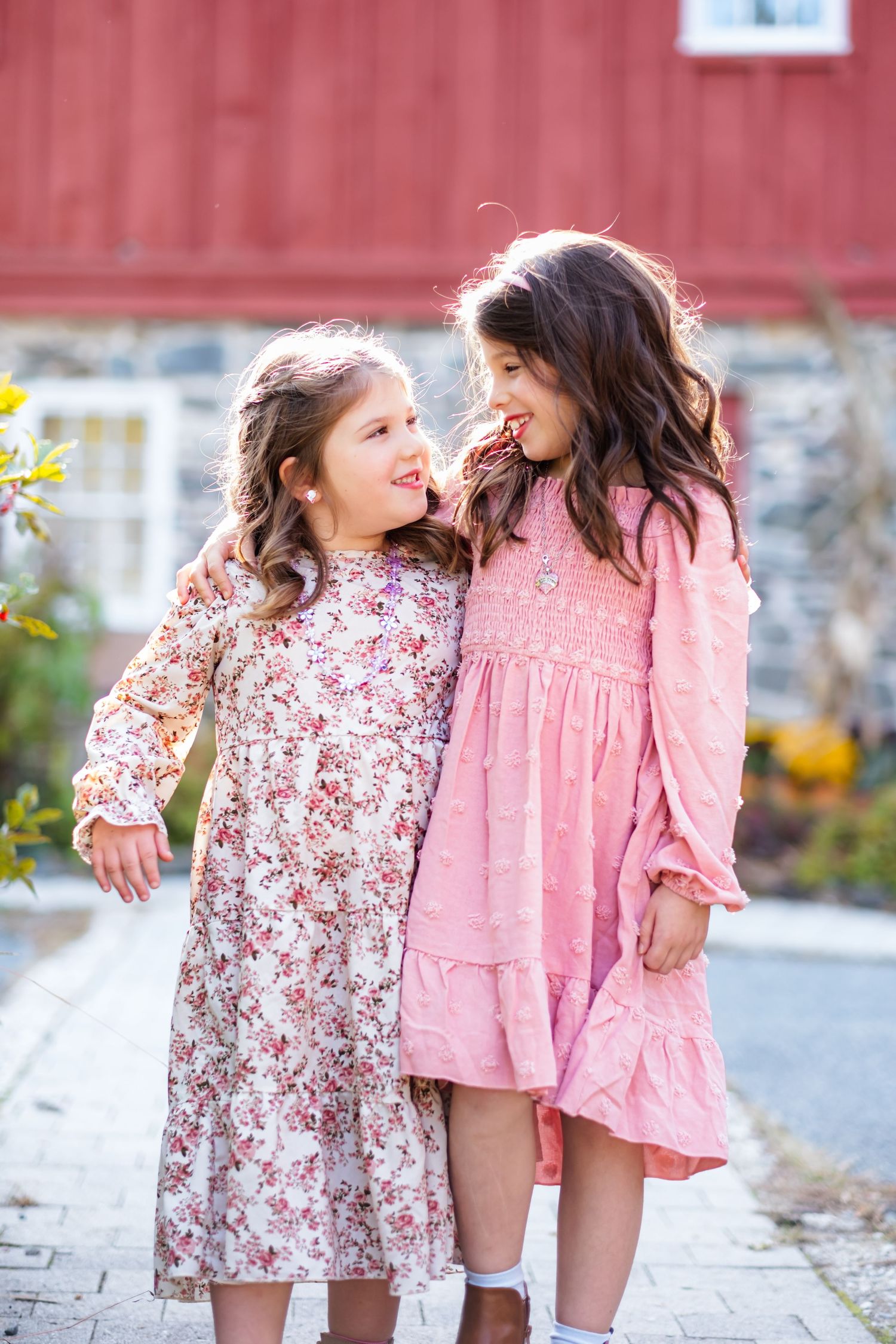 Two young girls in floral and pink dresses smiling and hugging outdoors near a red barn.
