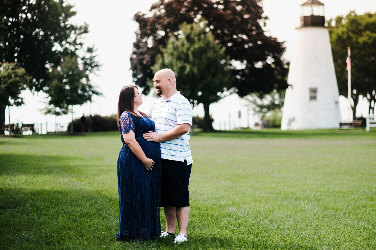 Pregnant couple standing on green lawn near historic lighthouse, woman in navy maternity gown, man in white polo shirt.