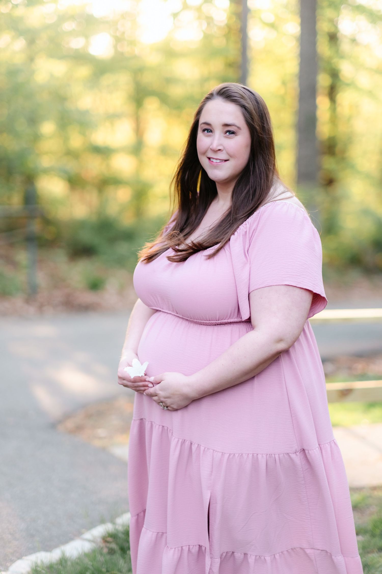 Smiling pregnant woman in pink maxi dress holding baby shoes outdoors during maternity shoot.
