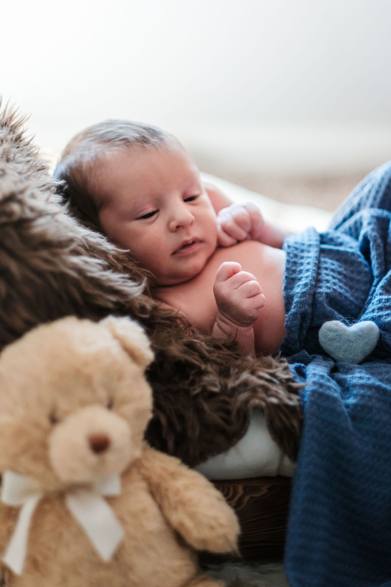 Newborn baby resting on fur blanket beside a teddy bear, wrapped in a soft blue knit blanket.