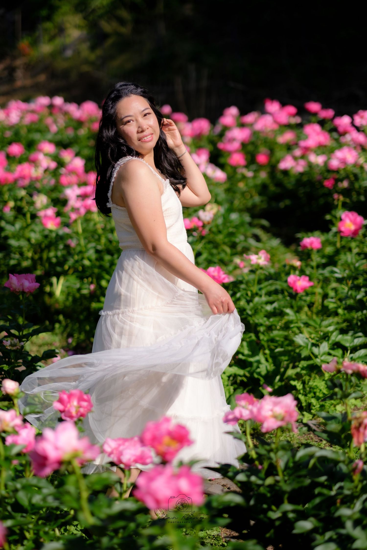 A bride in a white gown poses gracefully among blooming pink roses in a lush garden.