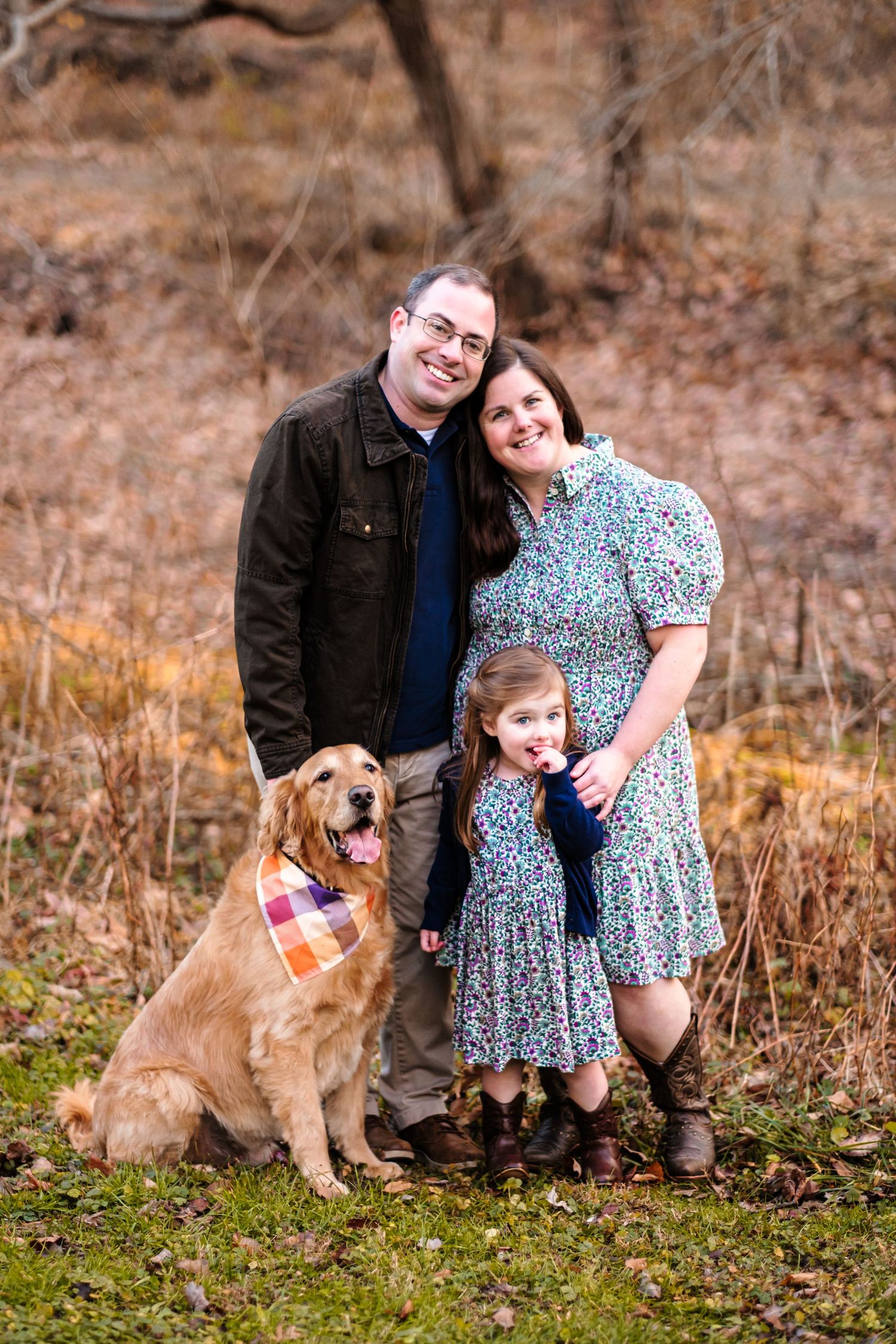 Family portrait with parents, young daughter, and golden retriever dog wearing plaid bandana, posing outdoors in autumn setting.