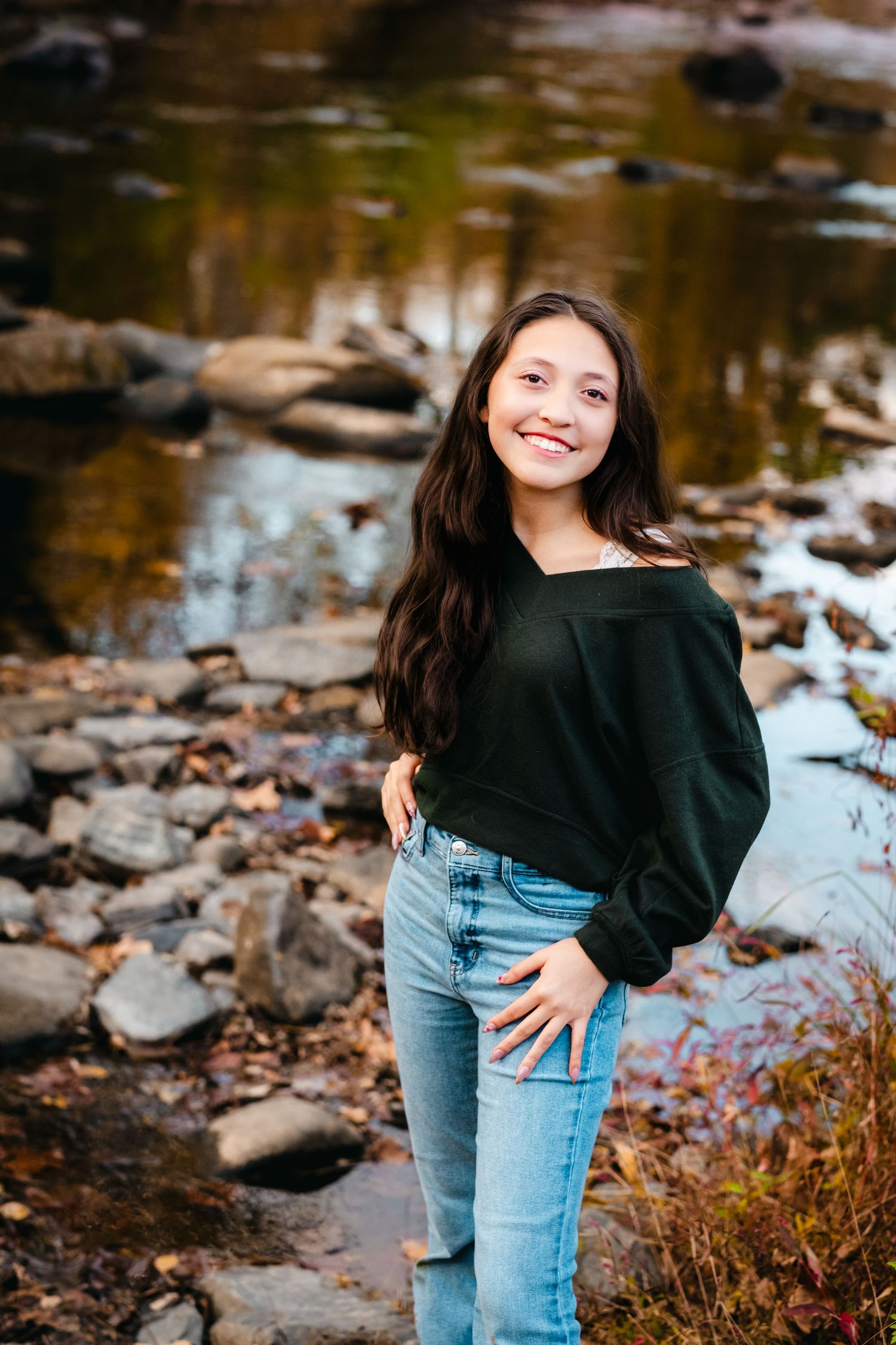 Smiling young woman in dark green off-shoulder top and jeans posing by a rocky stream in autumn.