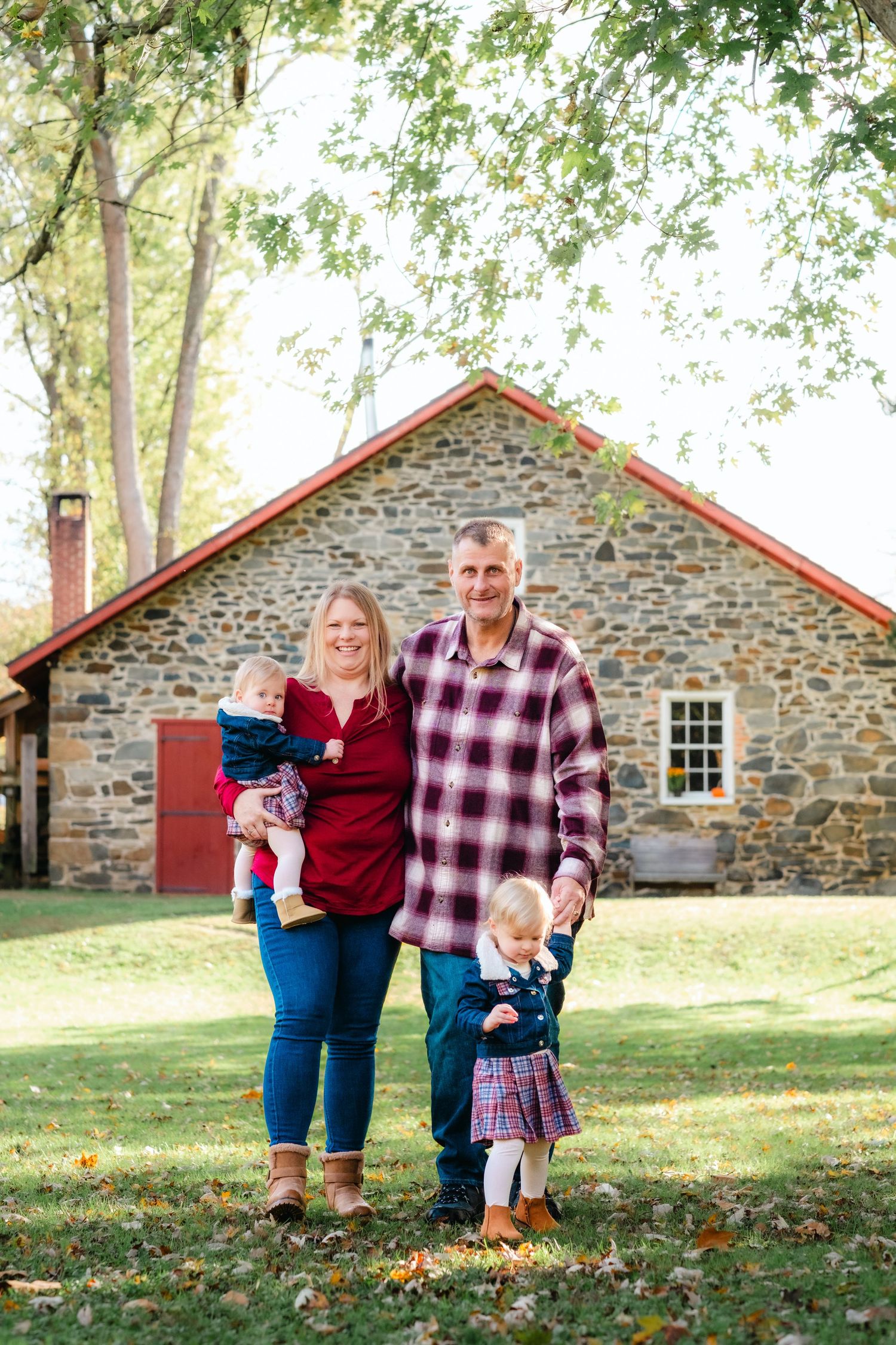 Family of four posing outdoors in front of a rustic stone building in autumn, parents holding young children.