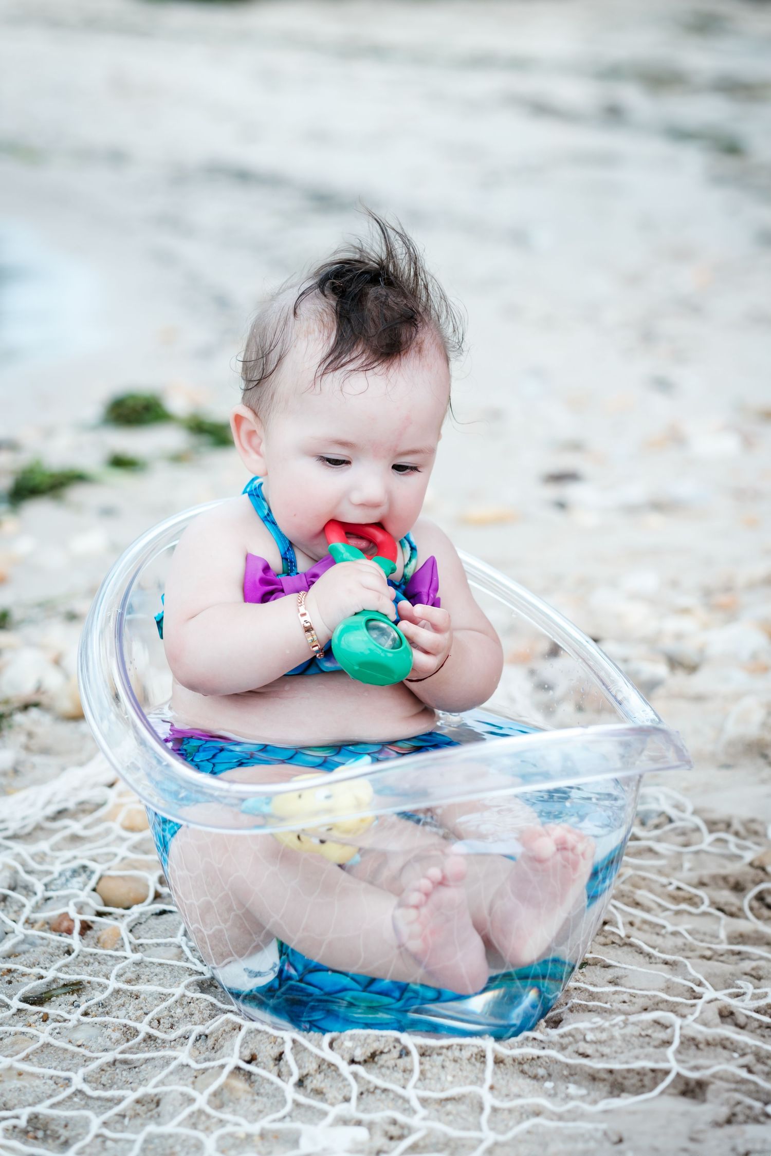 Baby girl sitting in a clear bowl on a sandy beach, chewing on a colorful toy while wearing a floral swimsuit.