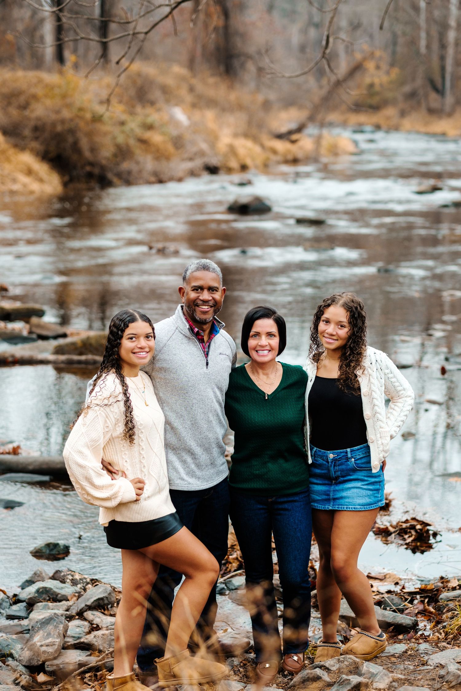 Family of four smiling together by a rocky stream in an autumn forest setting.