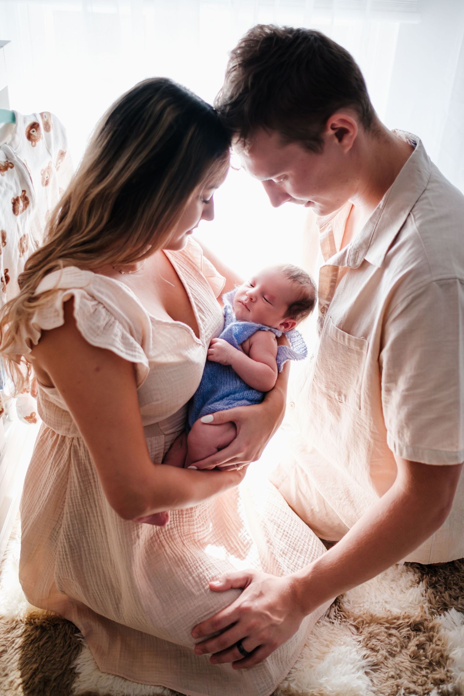 Young parents lovingly gazing at their newborn baby dressed in blue in a sunlit nursery room.