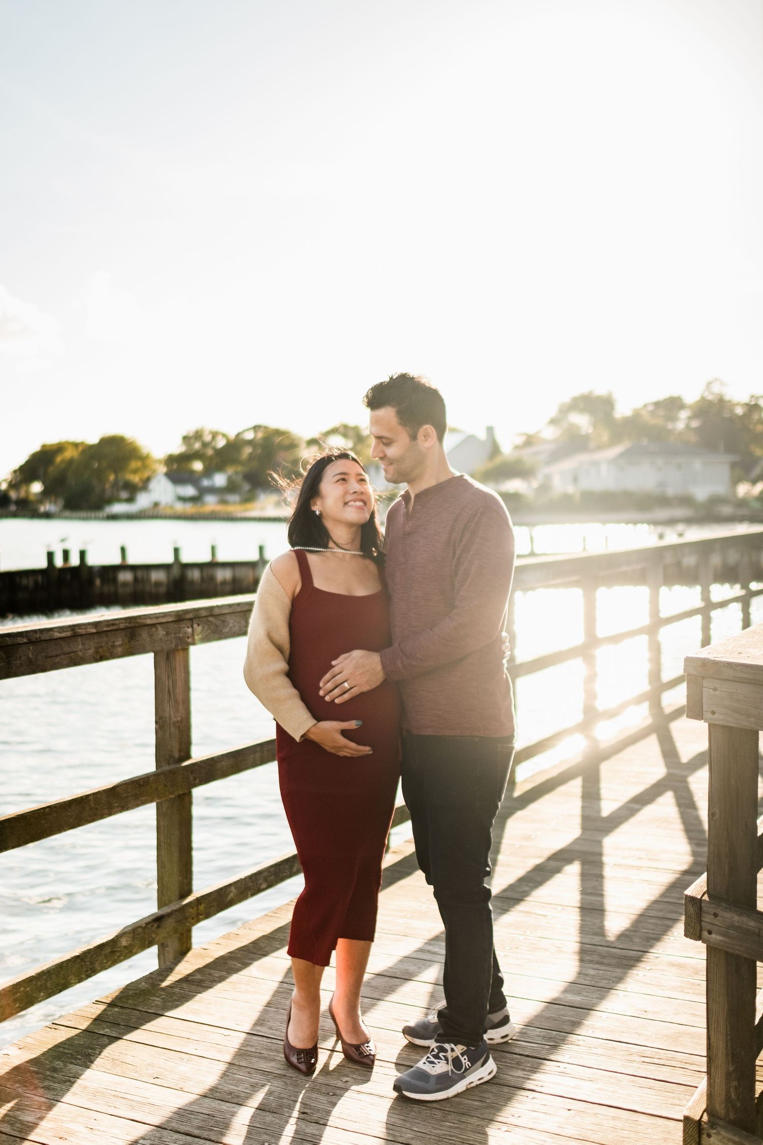 Pregnant couple embracing on a waterfront pier at golden hour, woman in burgundy dress with hands on belly.
